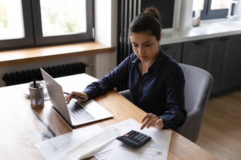A woman sits at a table using a laptop and calculator, surrounded by papers and a pen holder, appearing focused while working in a modern home setting with large windows.