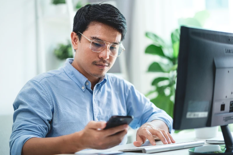 A man wearing glasses is sitting at a desk with a computer and a cell phone in his hand